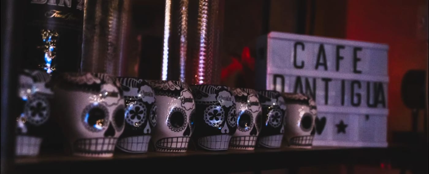 A lit Cafe D'Antigua sign among skull decorations on a bar shelf